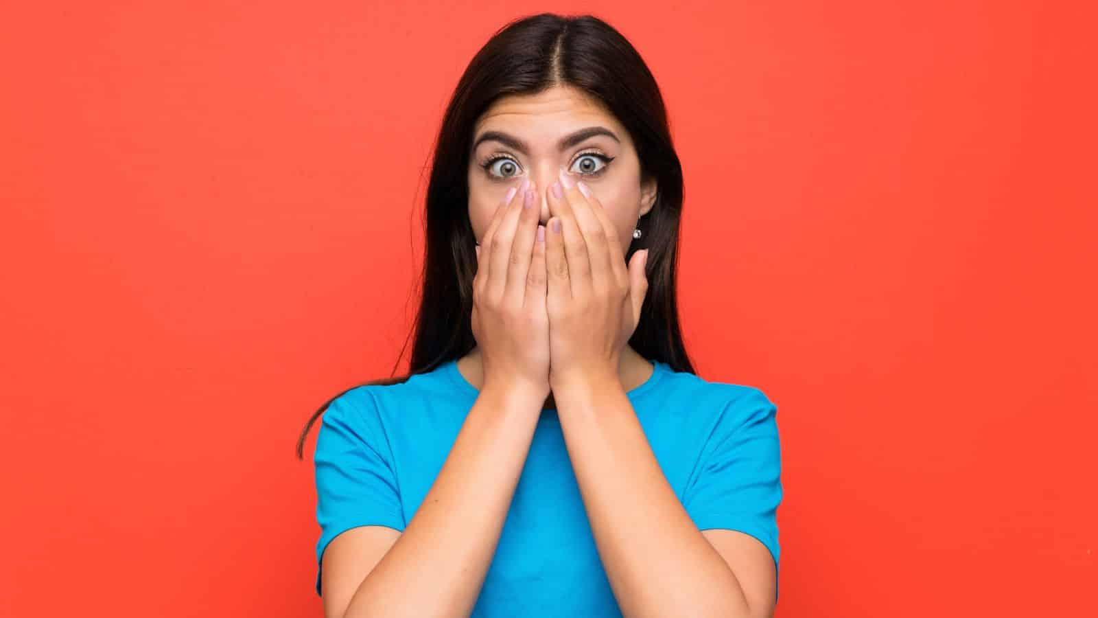 Woman in a blue shirt covering her mouth with both hands, looking surprised, against a red background.