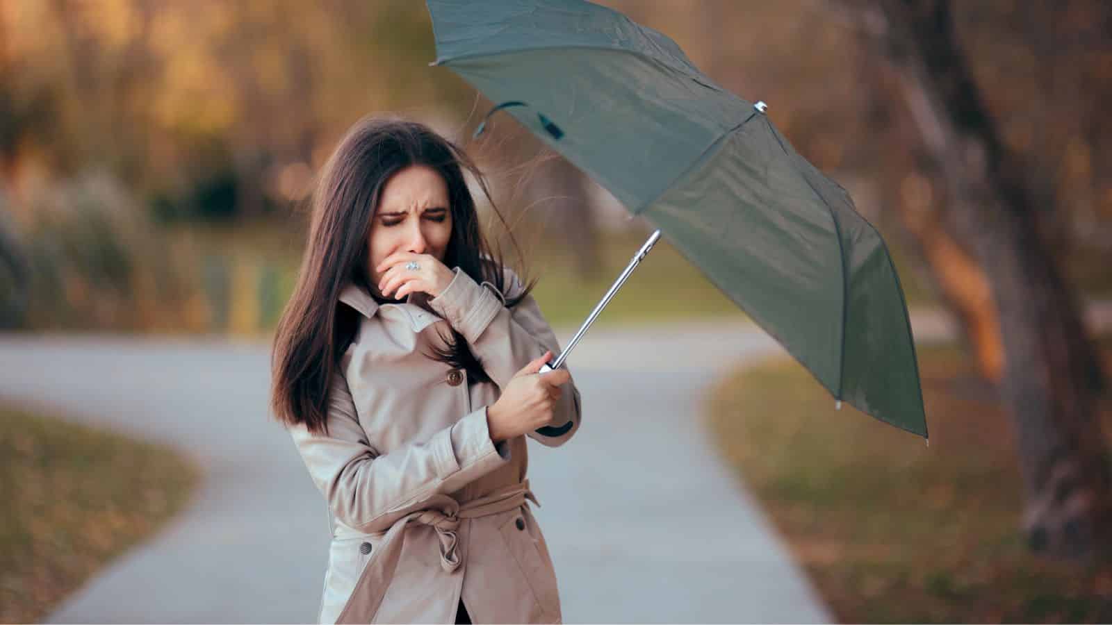 Woman in a coat struggling with an inside-out umbrella on a windy day in a park.