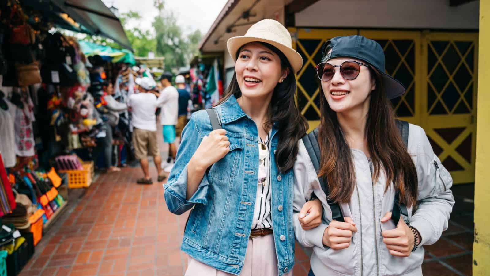 Two young women shopping outdoors, smiling and walking arm in arm, surrounded by market stalls.