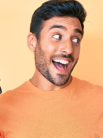A man with dark hair and a beard, wearing an orange t-shirt, smiles and gestures with his thumb while holding a closed brown wallet, standing in front of a light orange background—perfect for representing budget travel.