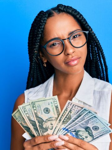 Woman with braids and glasses holding cash, looking skeptical, standing against a bright blue background.