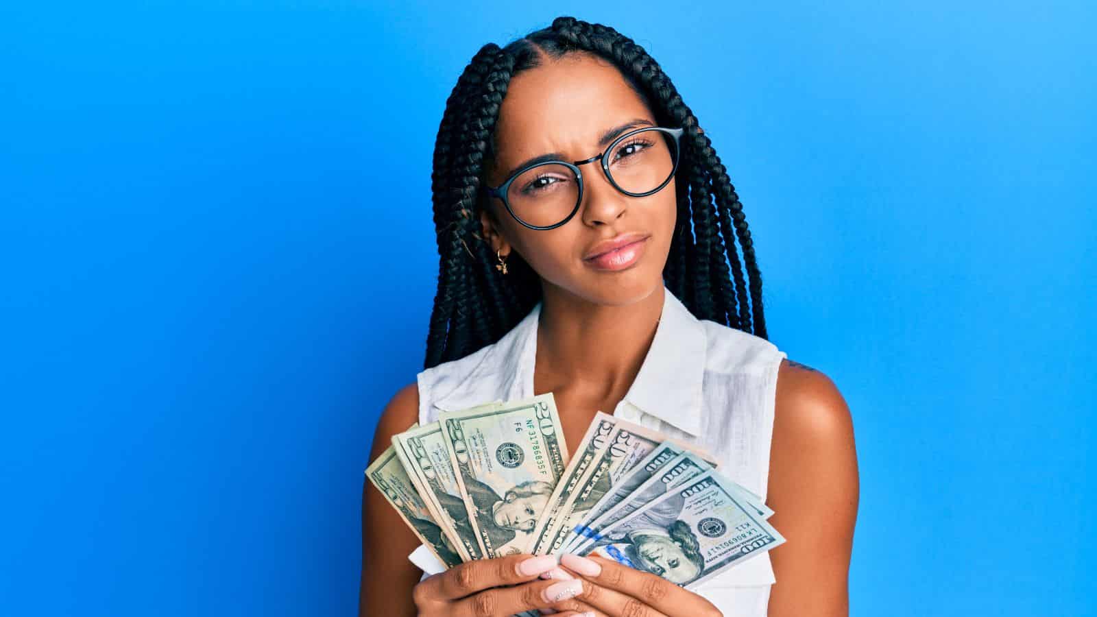 Woman with braids and glasses holding cash, looking skeptical, standing against a bright blue background.