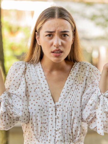 A woman outdoors shrugs with raised hands, looking confused or uncertain.