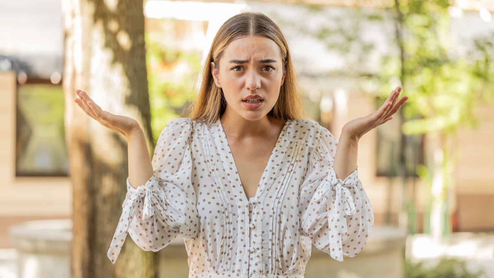 A woman outdoors shrugs with raised hands, looking confused or uncertain.