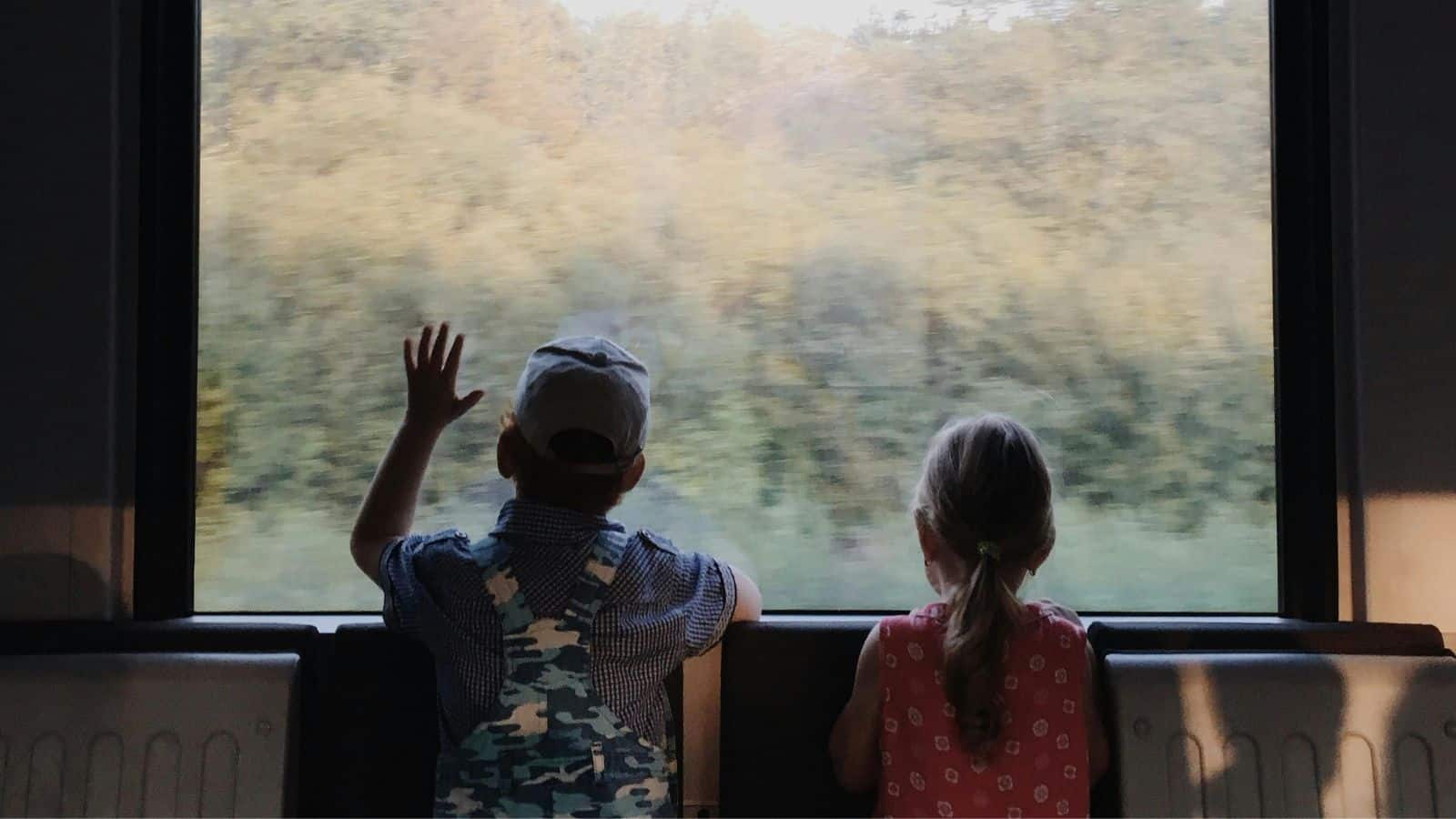 Two young children, one in overalls and a cap, the other in a pink dress with a ponytail, sit side by side on a train ride, gazing out the window at blurred trees—a heartwarming moment of train travel.