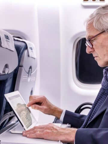 Older man in a suit uses a tablet while seated on an airplane, next to a window.