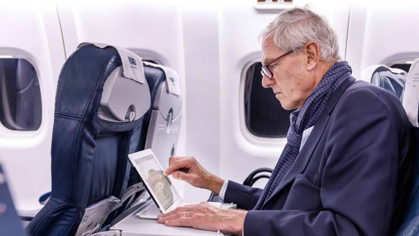 Older man in a suit uses a tablet while seated on an airplane, next to a window.