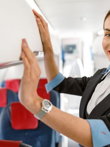 A smiling flight attendant arranges items in the overhead bin inside an airplane cabin.