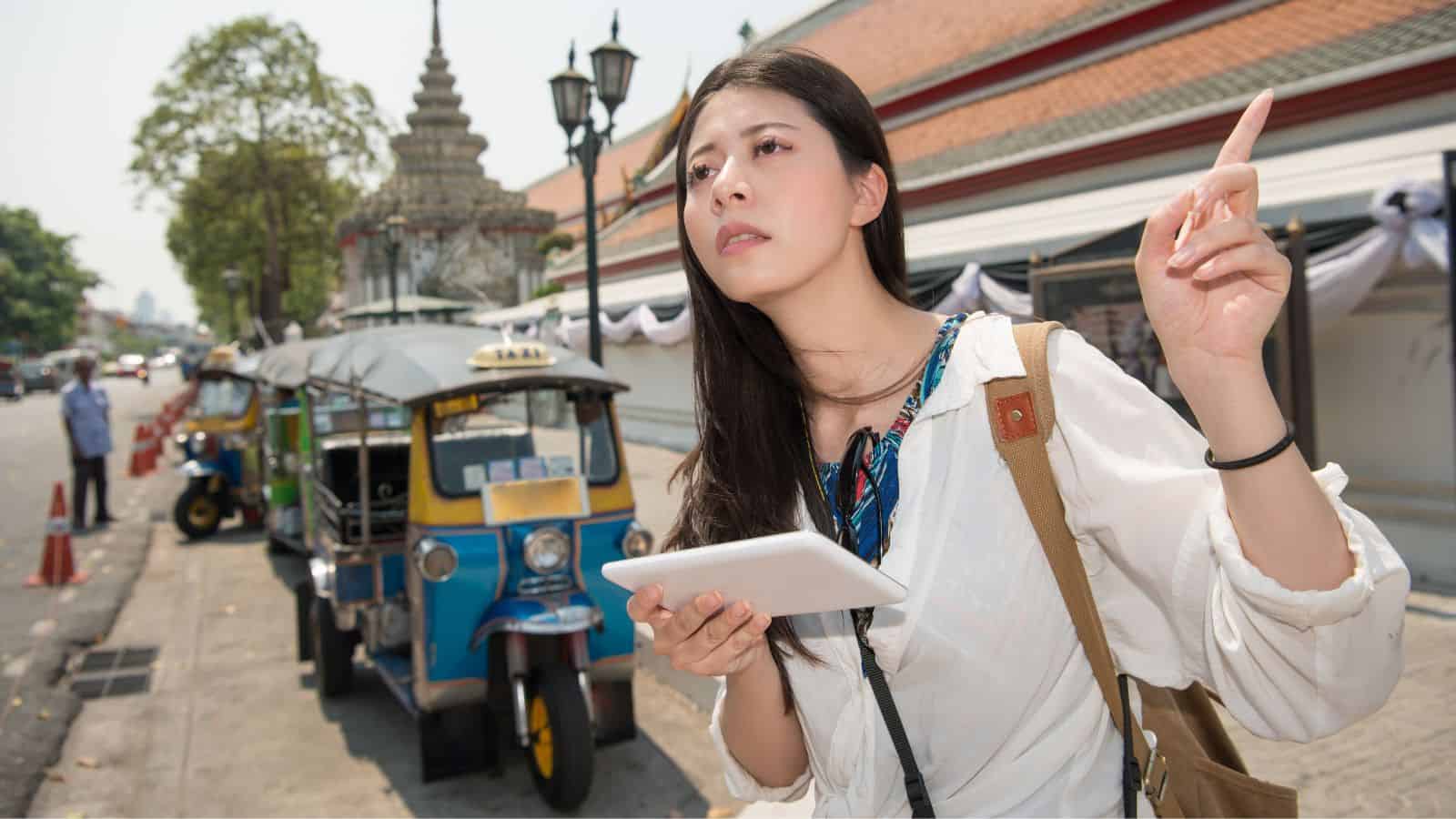 A woman holding a tablet points upward on a city street near tuk-tuks and a temple-like building.