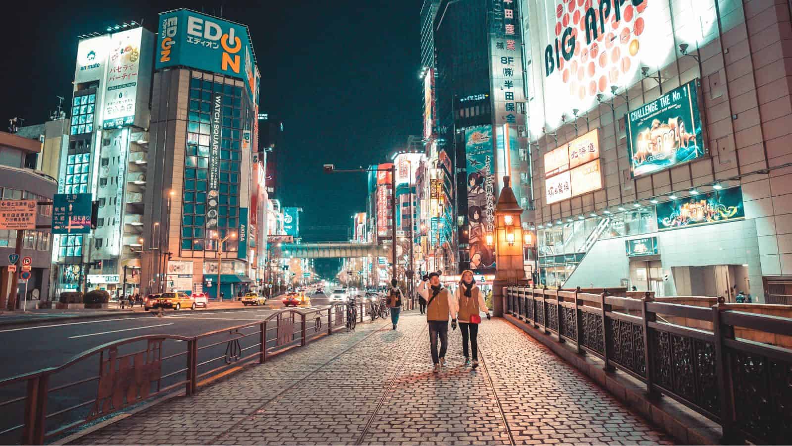 Two people walk on a lit street in a city at night, surrounded by tall buildings with bright neon signs.