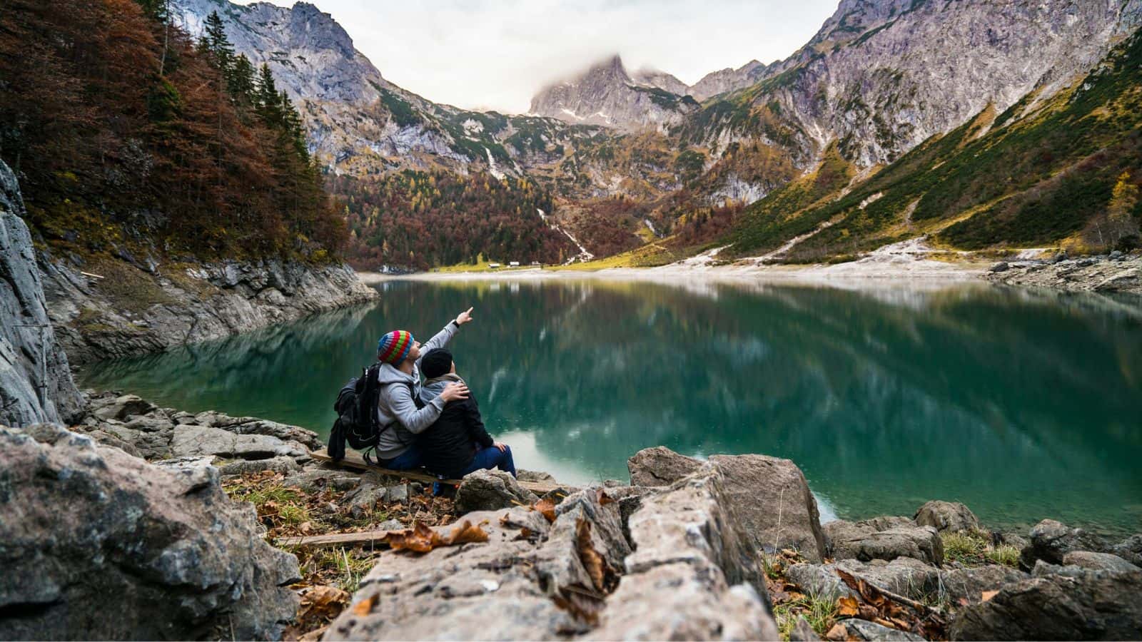 A couple sits by a mountain lake, one pointing toward the mountains, surrounded by rocky and forested terrain.