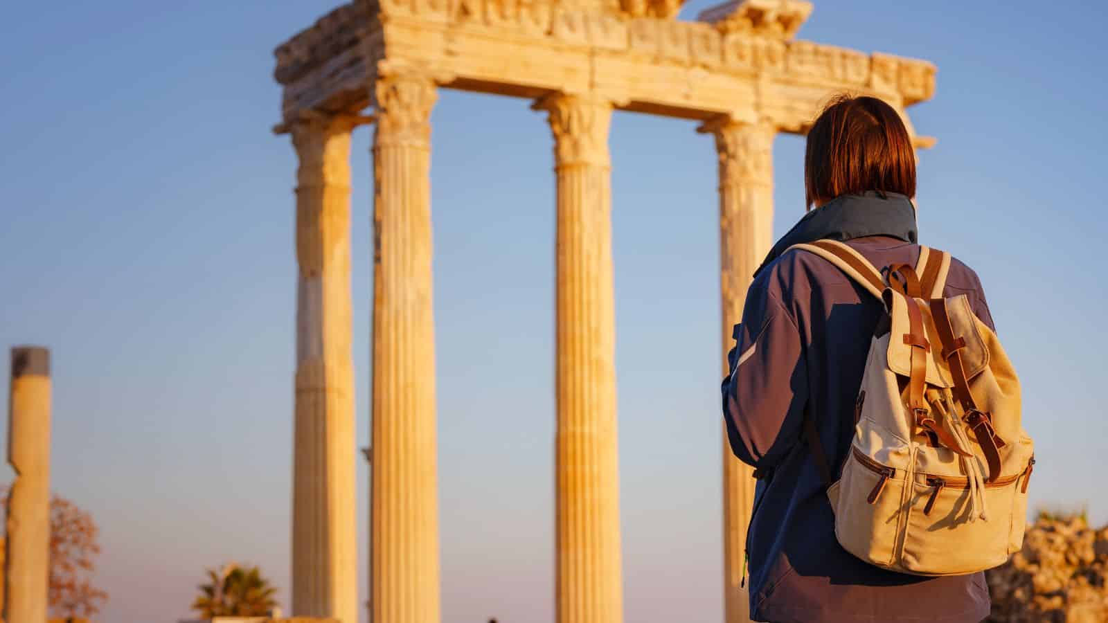 Person with a backpack stands in front of ancient Greek columns at sunset.