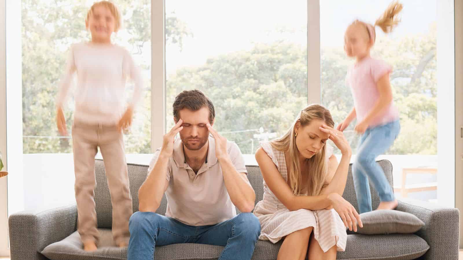 A stressed couple sits on a couch while two kids jump and play energetically in the background.