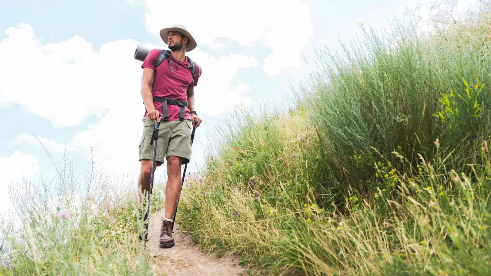 A person wearing a hat, maroon shirt, and green shorts hikes on a narrow dirt trail surrounded by tall grass and shrubs under a partly cloudy sky, carrying a backpack with a rolled mat.