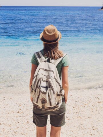 A traveler wearing a hat, green shirt, shorts, and a backpack stands on a pebbled beach facing the blue sea, rocky cliffs in the distance—a scene that captures the spirit of vacation trends and adventure.