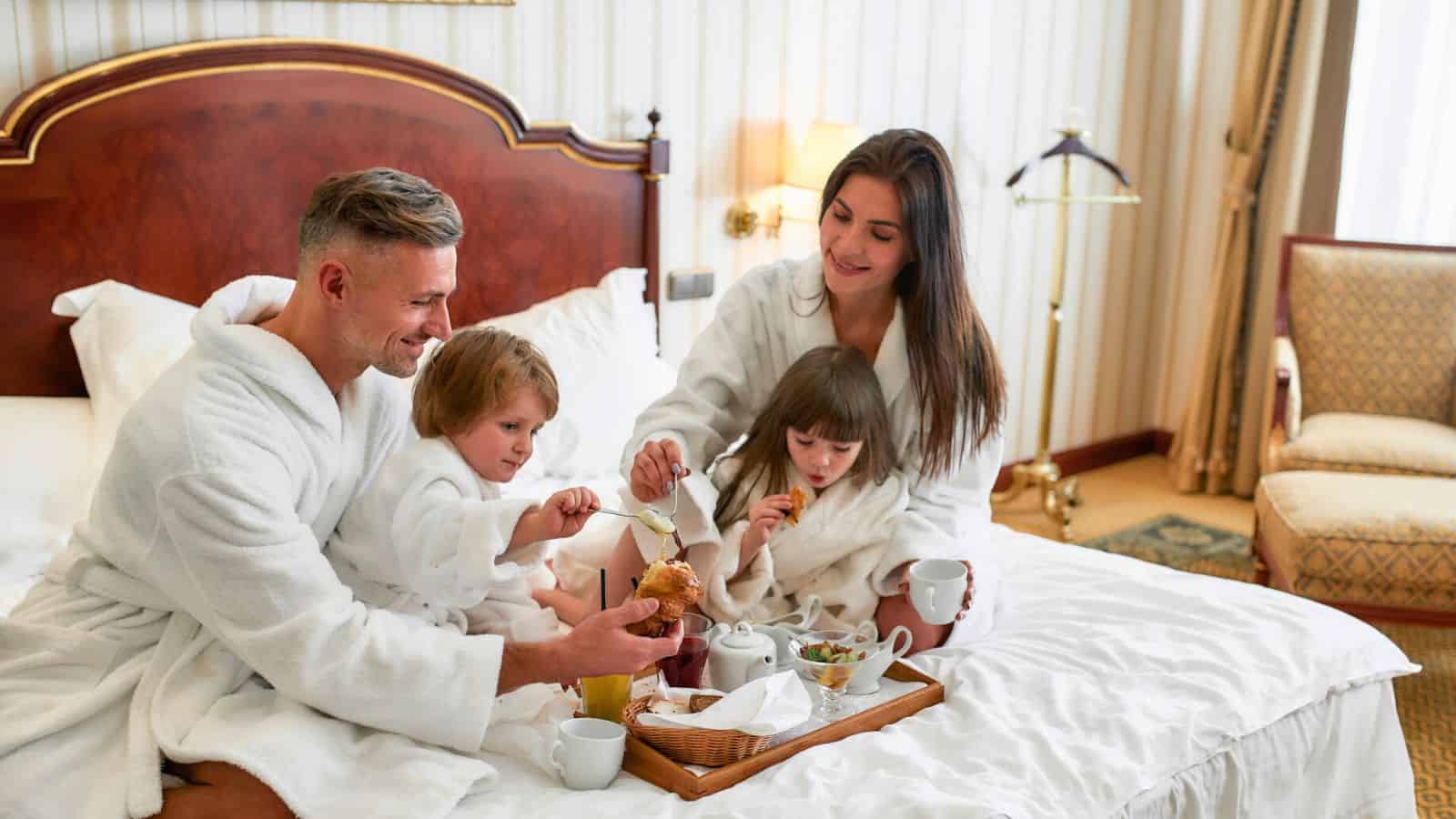 A family in white robes enjoys breakfast together on a bed in a cozy hotel room.