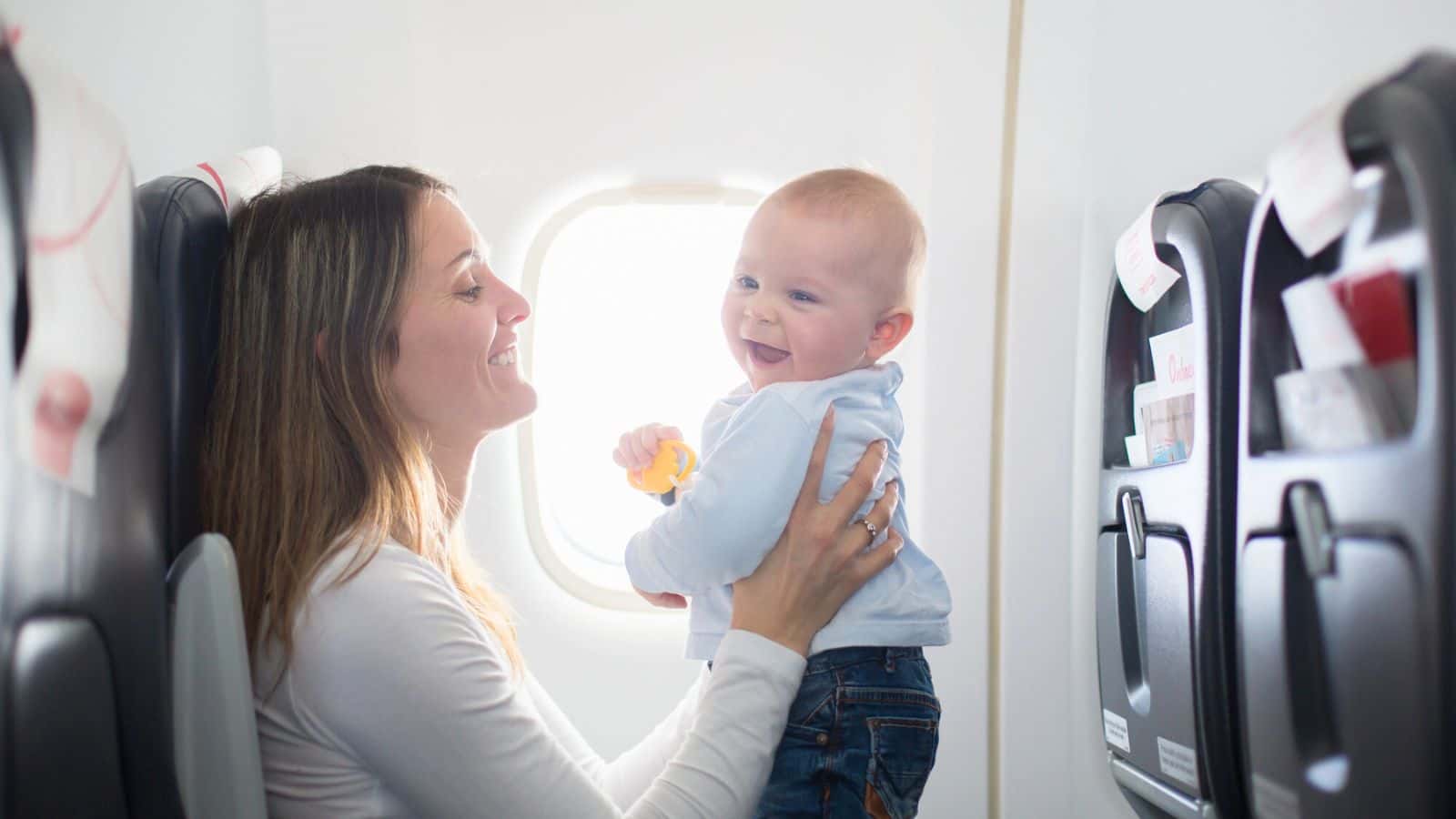 A woman holding a smiling baby near an airplane window, both seated in an aircraft cabin.