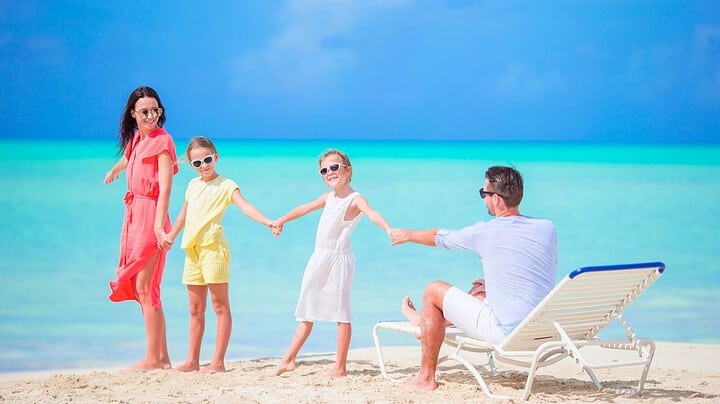 Family of four holding hands, smiling on a tropical beach with turquoise water and blue sky.
