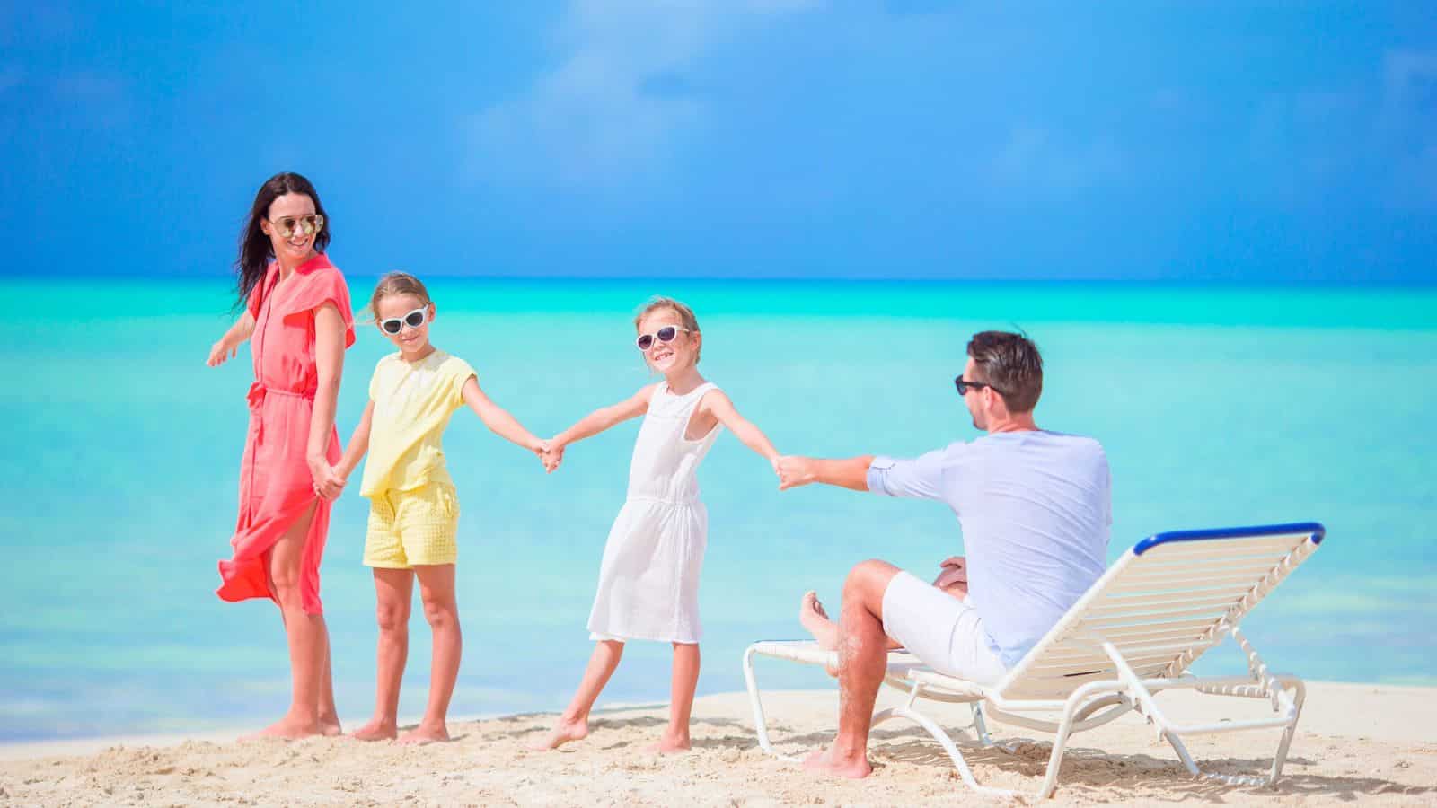 Family of four holding hands, smiling on a tropical beach with turquoise water and blue sky.