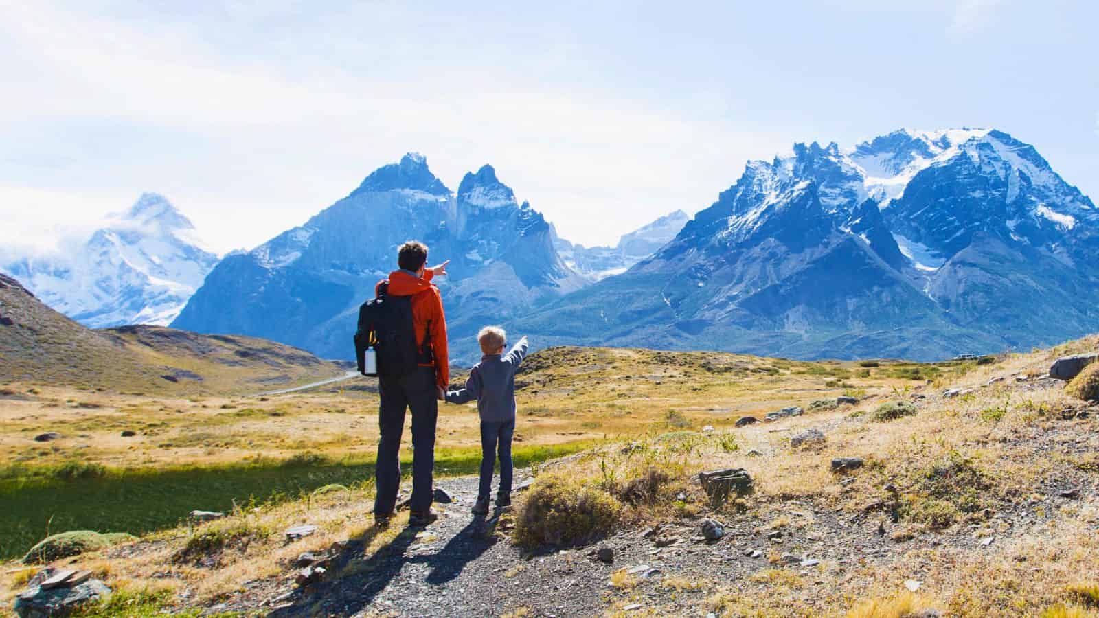 Adult and child holding hands, looking at distant snow-capped mountains in a vast, grassy landscape.