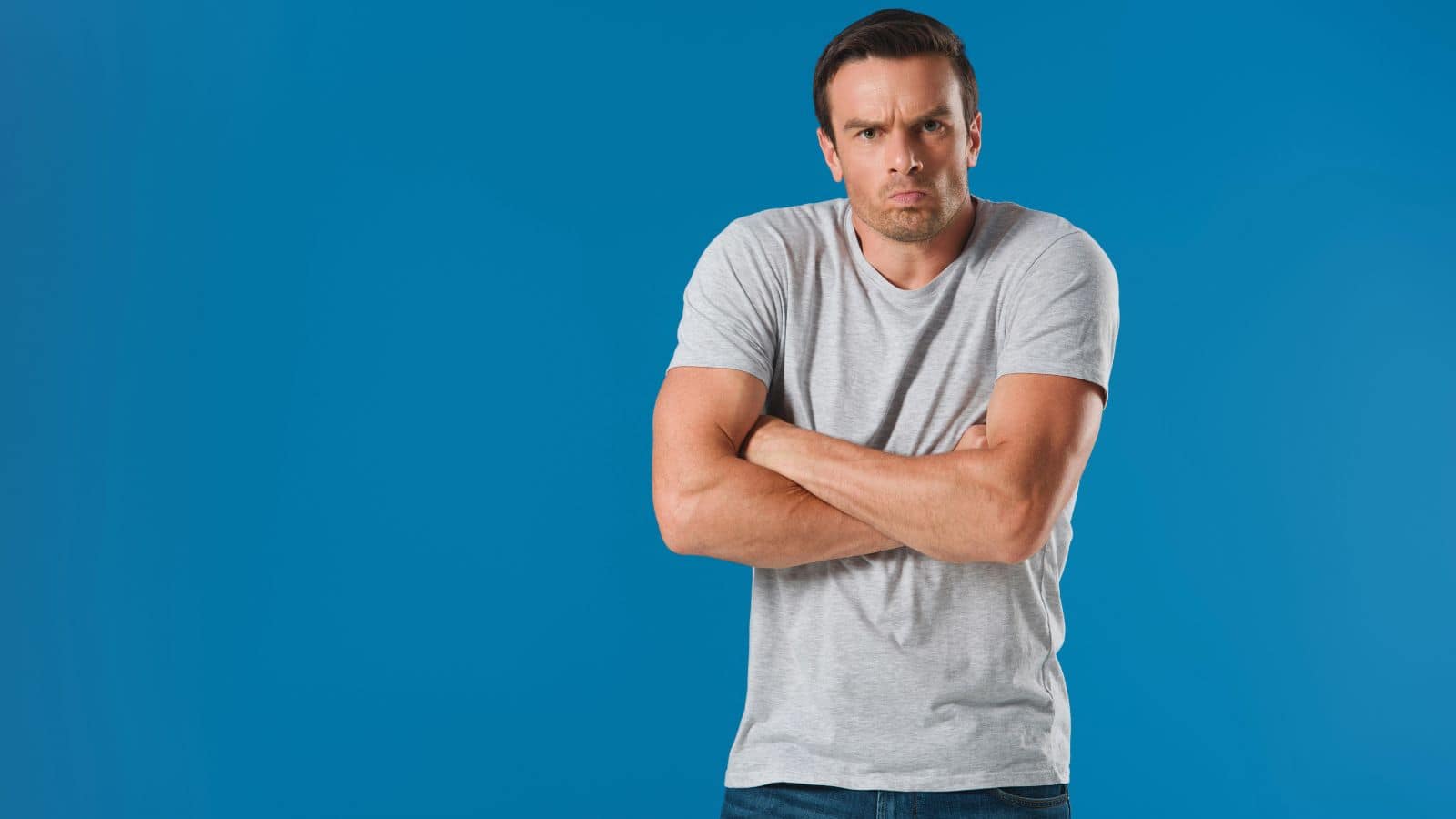 A man in a grey t-shirt and jeans stands against a blue background with his arms crossed, looking directly at the camera with a serious, almost jealous expression—perhaps thinking about U.S. things that are admired in other countries.