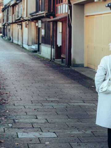 A woman with long brown hair wearing a white coat and carrying a white bag walks down a quiet stone-paved street lined with traditional wooden buildings and greenery—a perfect scene for solo travel enthusiasts exploring unique travel destinations.