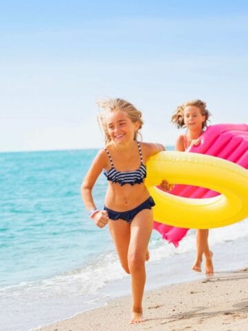 Three young children run along a sandy beach near the water. One carries a yellow inner tube, another holds a pink inflatable float, and the third carries a blue pool noodle. The sky is clear and sunny.