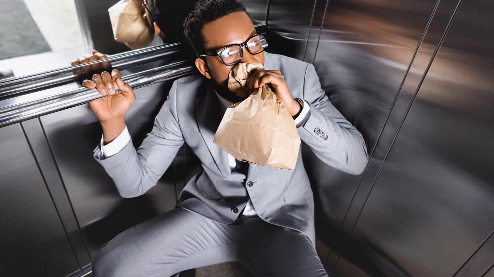 Man in a suit sitting in an elevator, breathing into a paper bag, looking anxious.