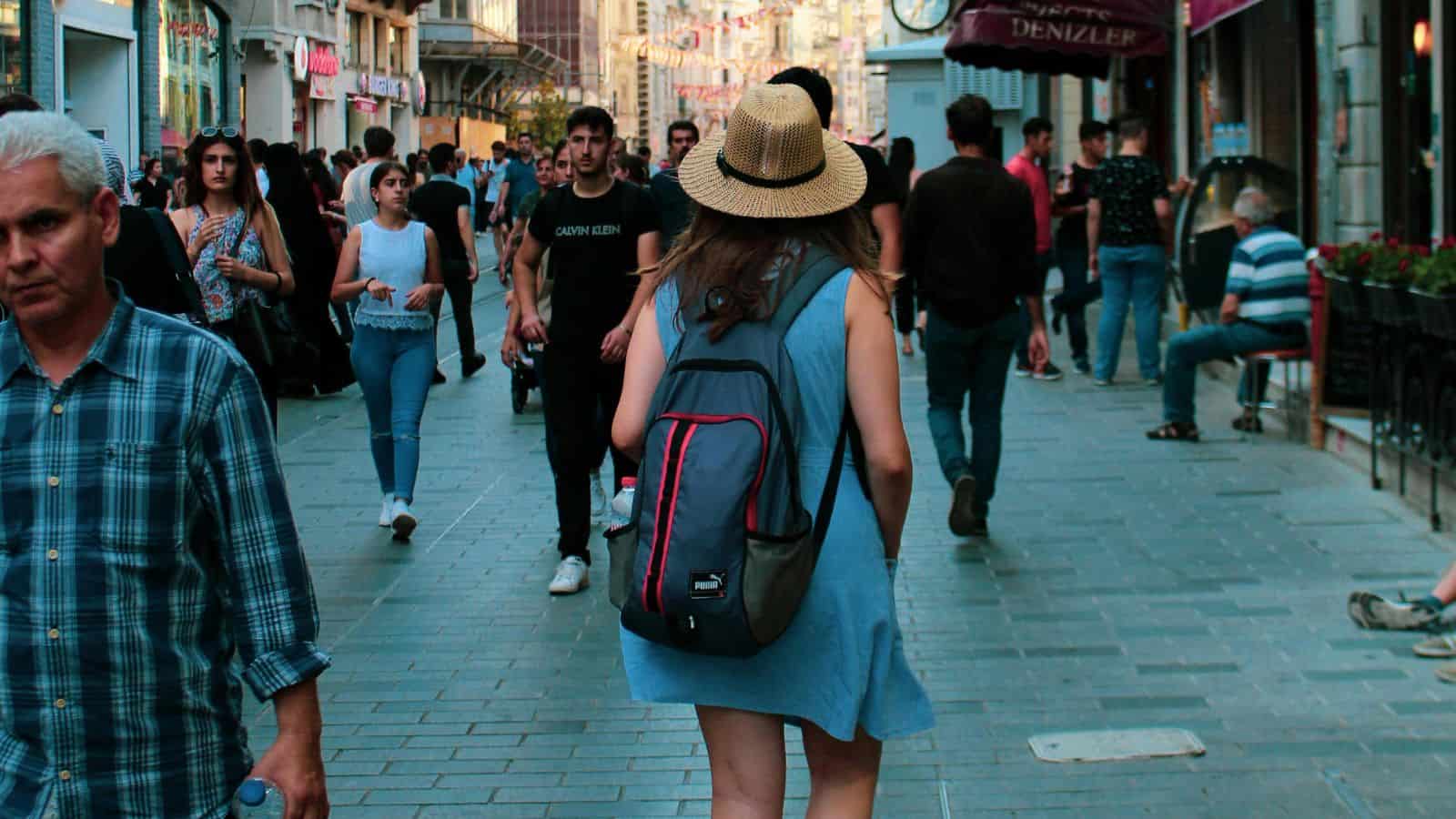 A woman wearing a straw hat, blue dress, and backpack walks down a crowded pedestrian street lined with shops&mdash;an inspiring scene for those seeking the best countries to visit on their first time abroad.