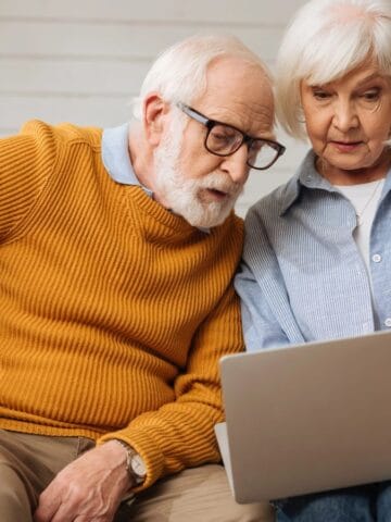 An older man in a mustard sweater and an older woman in a light blue shirt sit on a couch, looking at a laptop screen together as they research the best countries for retirement with focused expressions.