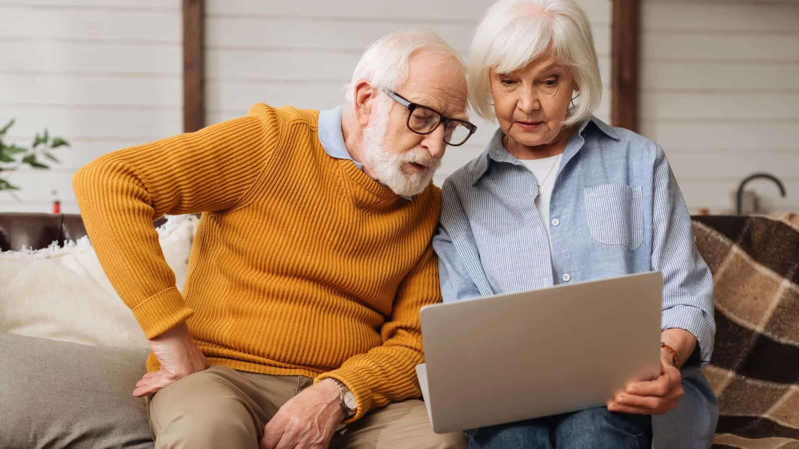 An older man in a mustard sweater and an older woman in a light blue shirt sit on a couch, looking at a laptop screen together as they research the best countries for retirement with focused expressions.