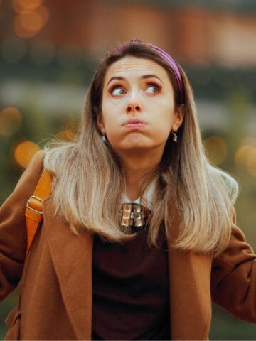 A woman with long hair wearing a brown coat and a headband stands outdoors with her arms raised, as if puzzled about travel preferences or which countries that prefer Canadians to visit. Blurred lights and greenery form the background.