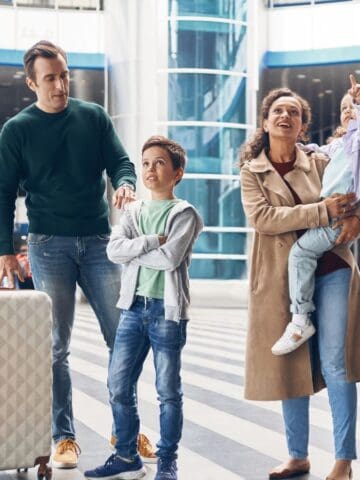 A family of four stands in an airport terminal. An adult man holds a suitcase, an adult woman holds a young girl, and a boy stands between them. The woman and girl are smiling and pointing upward.