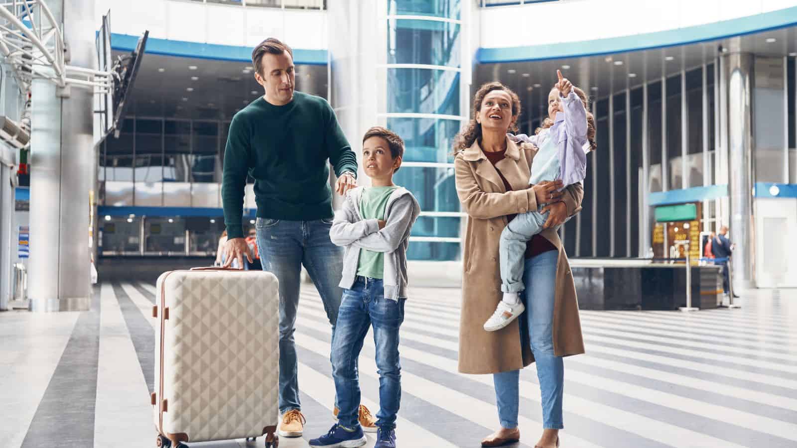 A family of four stands in an airport terminal. An adult man holds a suitcase, an adult woman holds a young girl, and a boy stands between them. The woman and girl are smiling and pointing upward.