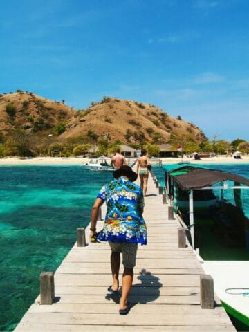A person in a blue floral shirt walks on a wooden pier toward a sandy island with hills and scattered trees, surrounded by clear turquoise water. A small boat is docked at the pier under a sunny blue sky.