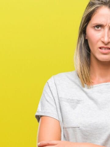 Woman with straight, shoulder-length hair wearing a light gray t-shirt stands against a yellow background, looking directly at the camera with a confused or frustrated expression.