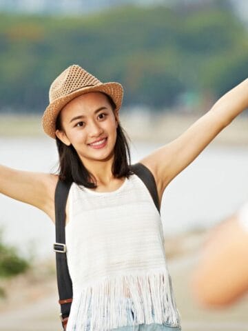 A young woman wearing a hat and backpack smiles with her arms outstretched while posing for a photo outdoors, with water and trees in the background. Another person is taking her picture.