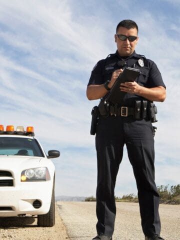 A police officer stands on the side of a road near a white police car with orange lights, possibly checking travel restrictions or special laws for tourists, with a clear sky and desert landscape in the background.