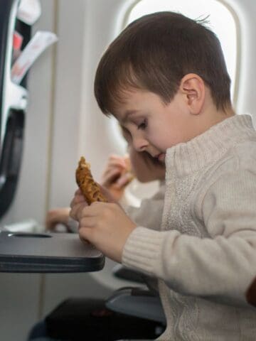 A young boy in a beige sweater sits in an airplane seat, leaning over a tray table while holding a piece of food. Another passenger is seated beside him. Bright daylight comes through the window.