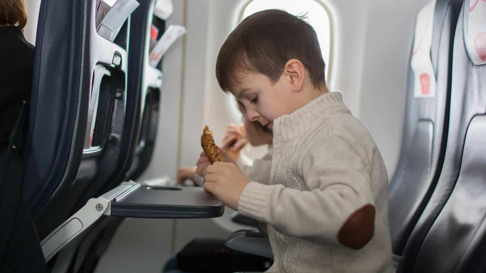 A young boy in a beige sweater sits in an airplane seat, leaning over a tray table while holding a piece of food. Another passenger is seated beside him. Bright daylight comes through the window.
