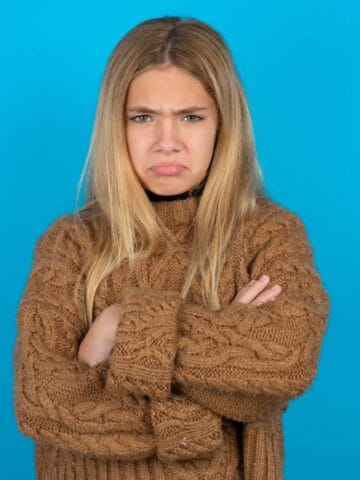 A young woman with long blonde hair stands against a solid blue background, wearing a brown knitted sweater and crossing her arms. She has a frown on her face and appears upset, perhaps annoyed by unwanted tourists during her travel.