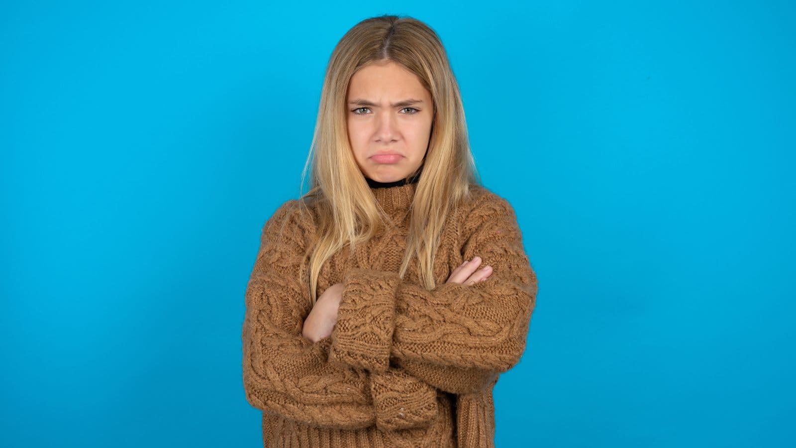 A young woman with long blonde hair stands against a solid blue background, wearing a brown knitted sweater and crossing her arms. She has a frown on her face and appears upset, perhaps annoyed by unwanted tourists during her travel.
