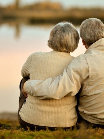 Two older adults with gray hair sit close together on the grass, facing a calm lake at sunset. They wear light-colored jackets and are seen from behind—an image of serenity for those exploring countries for retirees or retirement visas.