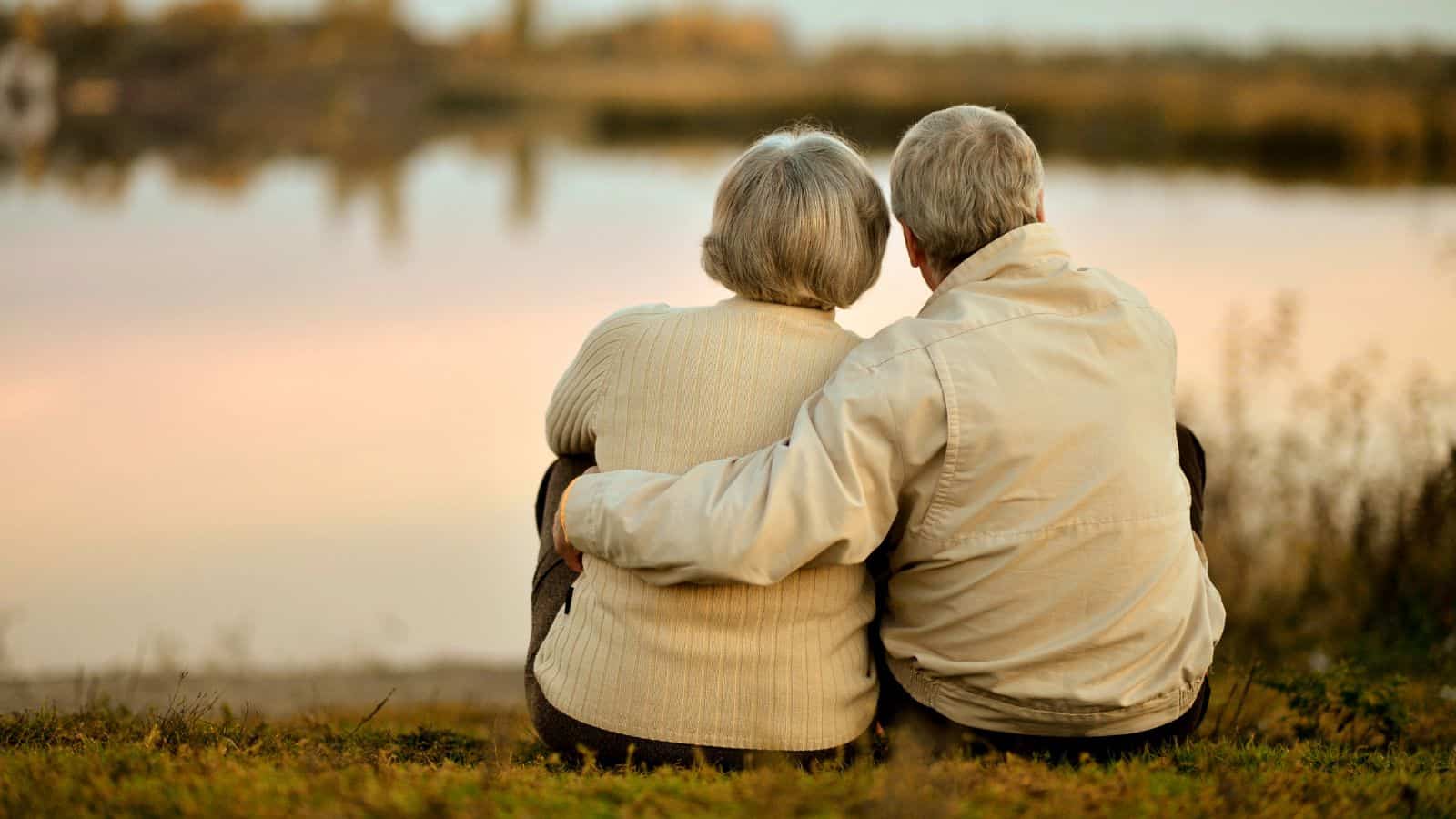 Two older adults with gray hair sit close together on the grass, facing a calm lake at sunset. They wear light-colored jackets and are seen from behind—an image of serenity for those exploring countries for retirees or retirement visas.
