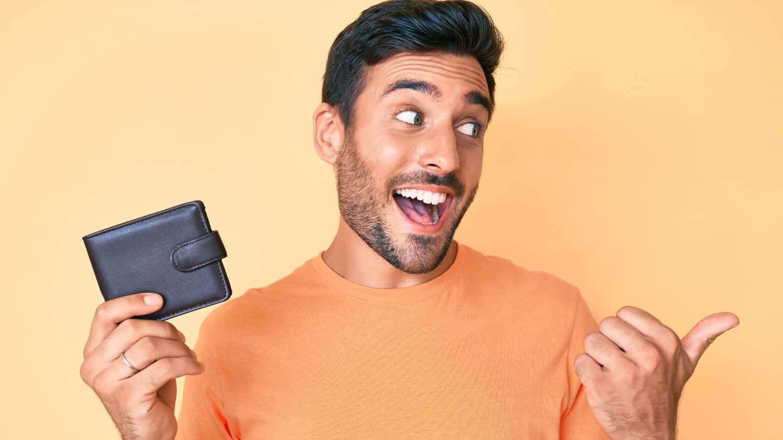 A man with dark hair and a beard, wearing an orange t-shirt, smiles and gestures with his thumb while holding a closed brown wallet, standing in front of a light orange background—perfect for representing budget travel.