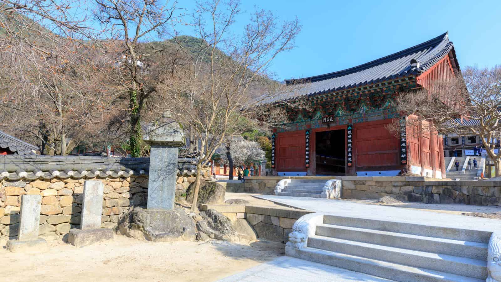Traditional Korean temple gate with ornate details, stone lantern, and bare trees under a clear blue sky.