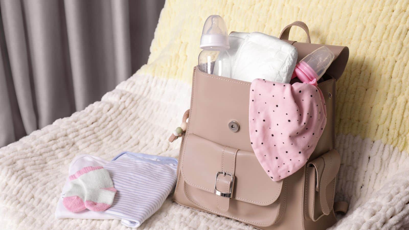 A beige diaper bag with baby bottles, diaper, and pink bib sits on a chair with baby clothes nearby.
