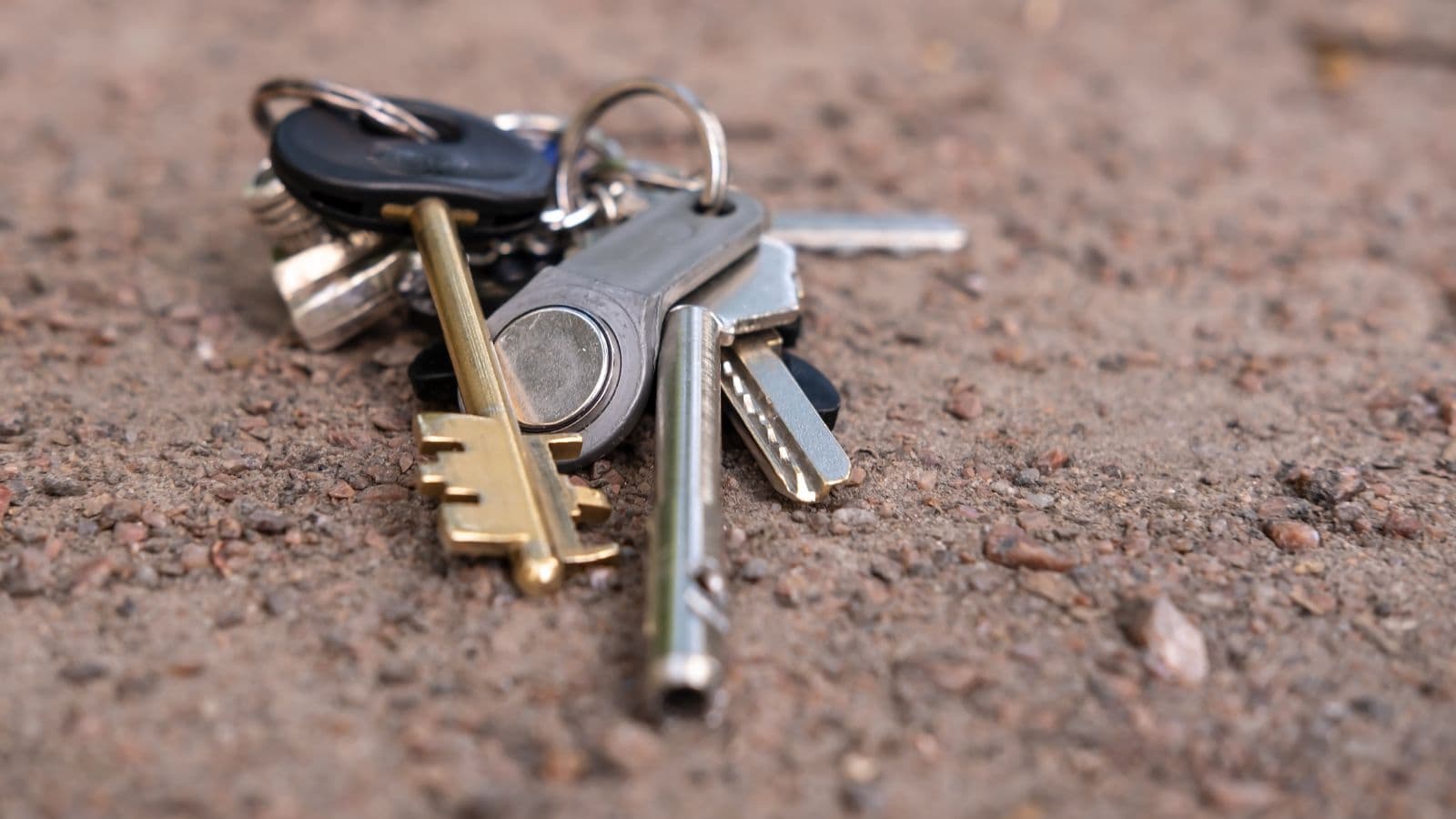 A set of various keys—commonly lost at U.S. airports—including a gold skeleton key and silver modern keys, attached to a keyring and lying on a rough, brown concrete surface.