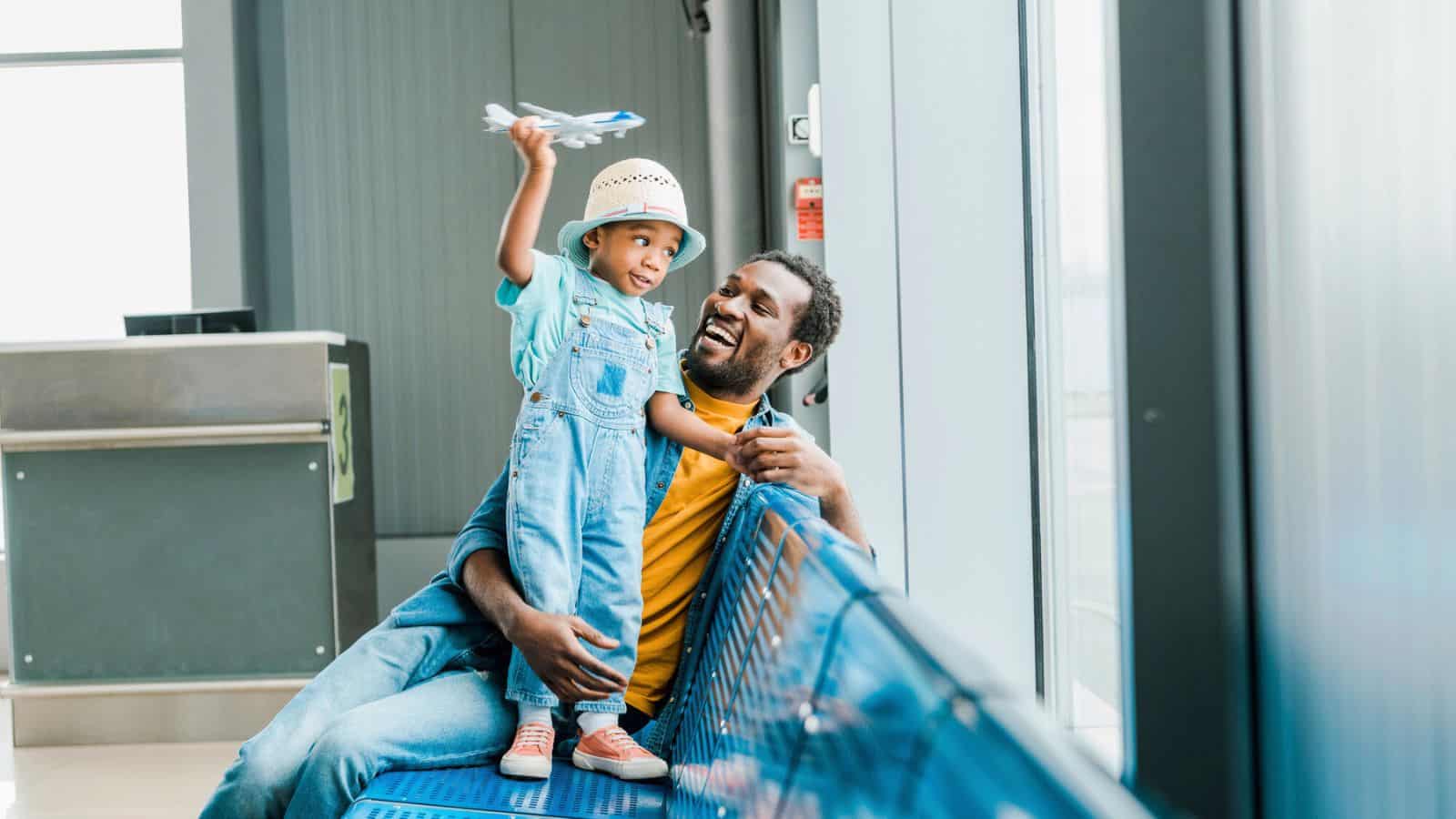 A child wearing overalls and a hat sits on an adult’s lap in an airport waiting area, holding a toy airplane, while the adult smiles up at the child. Both are sitting on a blue metal bench near large windows.