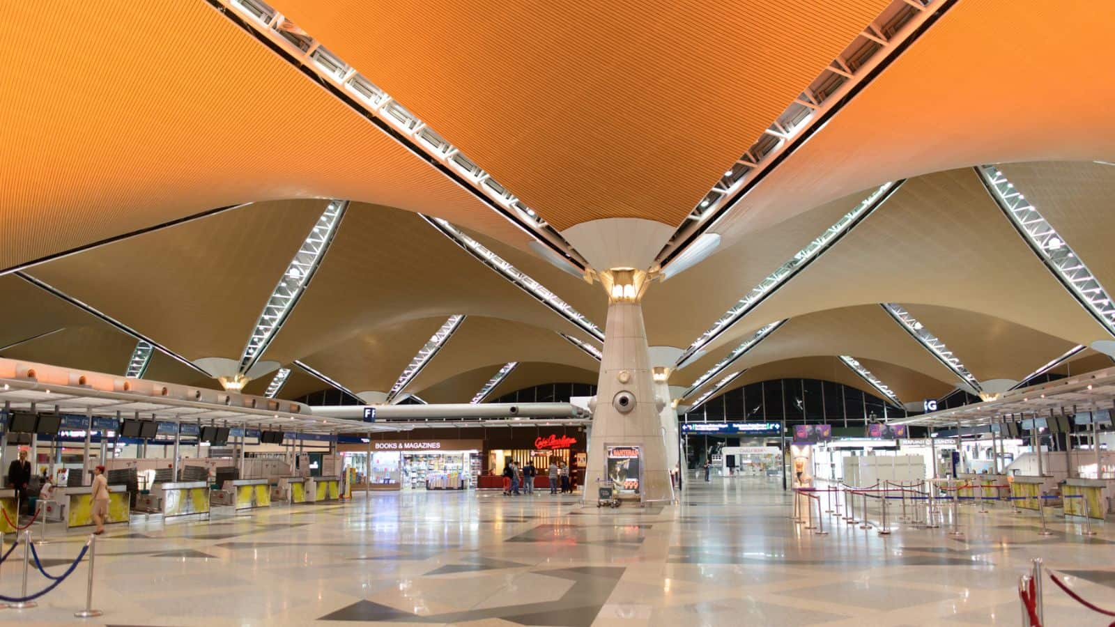 Spacious airport terminal with high, geometric orange and beige ceiling, check-in counters on either side, shops in the center, and polished floors. The area is well-lit and mostly empty.